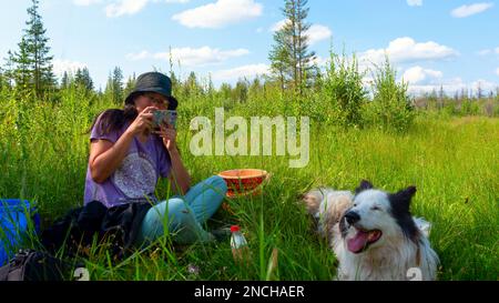 Ein Mädchen mit Hut, das auf dem grünen Gras sitzt, macht tagsüber ein Foto am Telefon mit einem weißen, fröhlichen Hund im Wald. Stockfoto