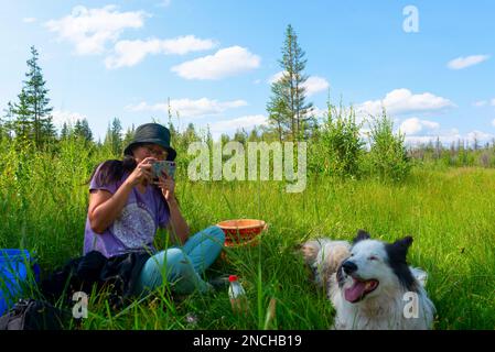Ein Mädchen mit Hut, das auf dem grünen Gras sitzt, macht ein Foto am Telefon mit einem weißen, fröhlichen Hund im Wald an einem sonnigen Tag. Stockfoto