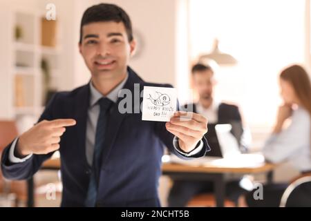 Ein junger Mann, der auf klebriges Papier zeigt, mit Text HAPPY DUMMYS TAG im Amt Stockfoto