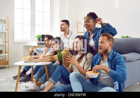 Eine multinationale Gruppe von Freunden von Fans versammelte sich vor dem Fernseher und saß auf dem Sofa im Zimmer am Couchtisch. Stockfoto