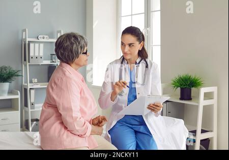 Seniorin beim Kontrolltermin mit einem jungen Arzt, der Symptome in flipboard schreibt. Stockfoto