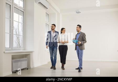 Ein gutaussehender, bärtiger männlicher makler im Anzug mit Dokumenten in seinen Händen rät einem jungen, glücklichen Paar, bevor er eine neue Wohnung kauft. Stockfoto