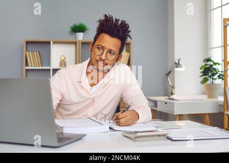 Ein junger indischer Geschäftsmann, der im Büro an einem Laptop arbeitet und auf einen Computerbildschirm schaut. Stockfoto