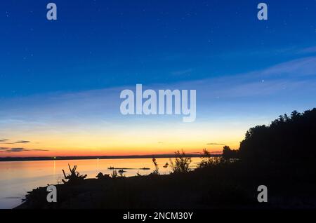 Hellblauer Sternenhimmel über dem leuchtend orangefarbenen Schein des Sonnenuntergangs über dem Fluss mit den Lichtern der Häuser auf einer Seite und einem Wald zum Strand Stockfoto