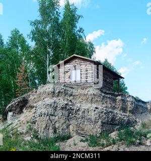 Kleines einsames Taigahaus Holz mit einem Fenster steht auf dem Hügel an der Klippe im Wald hinter den Birkenbäumen unter blauem Himmel mit Wolken. Stockfoto