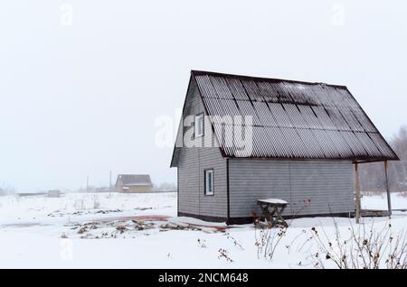 Privates rustikales 2-stöckiges Haus vom Nebengebäude ohne Zaun in einem Feld im Winter im Schnee und im Schneesturm im Hintergrund der Hütte Stockfoto