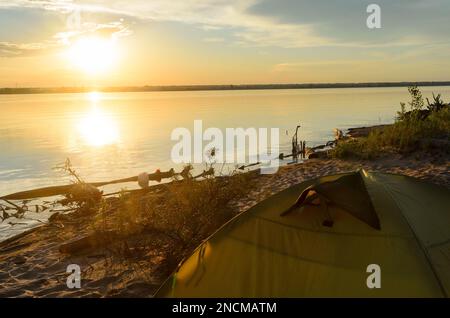 Top Touristenzelte, stehen am Sandstrand der Strandbäume, vor dem Hintergrund des hellen Sonnenuntergangs auf dem Fluss mit Reflexion im Wasser. Stockfoto