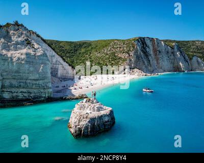 4K Drohnenaufnahme eines Paares, das mit einem Boot an einem Felsen im Wasser von Fteri Beach, Kefalonia, Cephalonia, Griechenland Händchen hält und Menschen sich sonnen Stockfoto