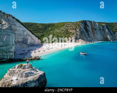 4K°-Aufnahme eines Paares auf einem Felsen am Fteri Beach, Kefalonia, Cephalonia, Griechenland mit Menschen, die sich sonnen, ein Boot, Grün, Kalksteinfelsen Stockfoto