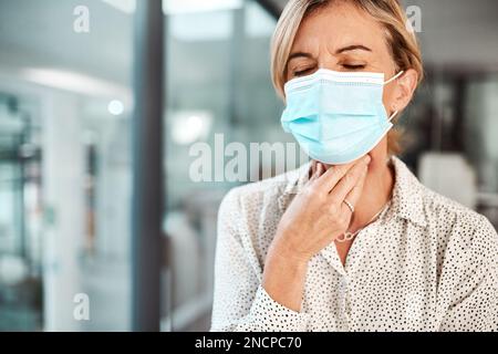 Ich brauche etwas, um dieses kratzende Gefühl zu lindern. Eine reife Geschäftsfrau, die eine Maske trägt und sich in einem Büro die Kehle reibt. Stockfoto