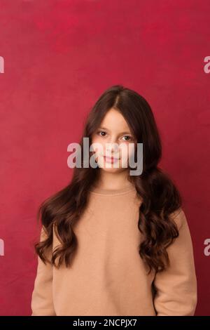 Schönes positives Mädchen mit langen lockigen Haaren, das auf pinkfarbenen Hintergrund in die Kamera schaut Stockfoto