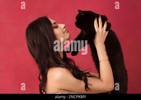 Eine schöne Frau, die eine schwarze Katze auf pinkfarbenem Hintergrund hält. Stockfoto