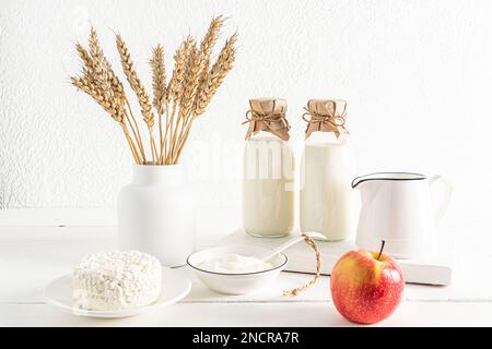 Vorderansicht von zwei Flaschen und einer Kanne Milch, Hüttenkäse, Sauerrahm in Schüsseln. Eine Vase mit Ähren aus Mais. Das Konzept des Shavuot-Urlaubs Stockfoto