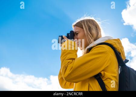 Wunderschöne Reiselin mit Rucksack fotografiert mit der Kamera vor blauem Himmel mit Wolken. Stockfoto