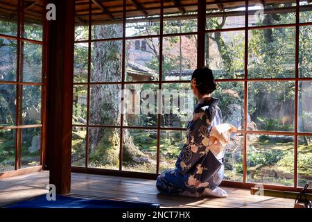 Eine junge Frau, die einen traditionellen japanischen Kimono trägt, sitzt am Fenster und blickt auf einen üppigen Garten Stockfoto