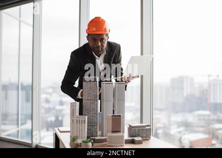 Fokussierter afrikanischer Inspektor, der mit einem Laptop ein Wolkenkratzermodell in einem architektonisch-modernen Büro inspiziert, mit Panoramablick auf die Stadt in einem architektonischen Büro. Ein hübscher Mann mit Anzug und Schutzhelm. Stockfoto