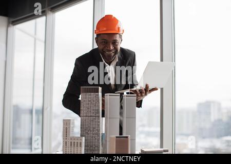 Fokussierter afrikanischer Inspektor, der mit einem Laptop ein Wolkenkratzermodell in einem architektonisch-modernen Büro inspiziert, mit Panoramablick auf die Stadt in einem architektonischen Büro. Ein hübscher Mann mit Anzug und Schutzhelm. Stockfoto