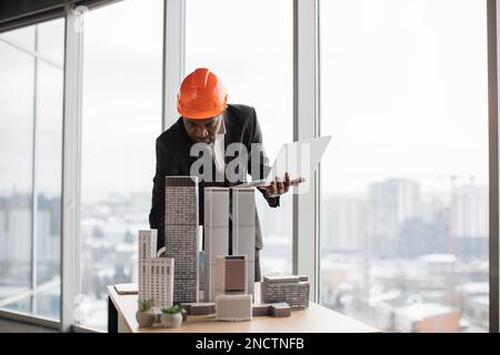Fokussierter afrikanischer Mann in schwarzem Anzug und orangefarbenem Schutzhelm, der ein digitales Notebook benutzt, um ein nachhaltiges 3D-Megalopolis-Modell auf dem Schreibtisch eines Büros mit Panoramafenstern und verschwommener Stadtlandschaft zu entwickeln Stockfoto