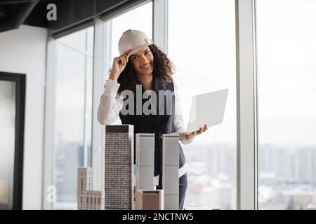 Lächelnde Architektin mit Laptop, die das Wolkenkratzermodell in einem architektonisch-modernen Büro inspiziert, mit Panoramablick auf die Stadt im architektonischen Büro. Attraktive Frau mit Anzug und Schutzhelm. Stockfoto