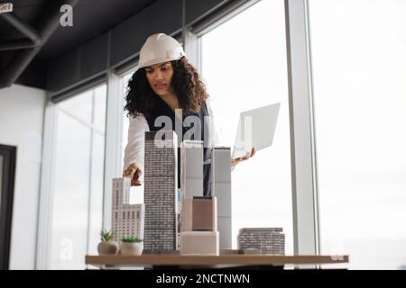 Junge Architektin, die mit einem Laptop ein Wolkenkratzermodell in einem architektonisch-modernen Büro inspiziert, mit Panoramablick auf die Stadt im architektonischen Büro. Attraktive Frau mit Anzug und Schutzhelm. Stockfoto