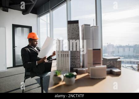 Büroeinrichtung mit Panoramablick auf die Stadt. Attraktiver Chefarchitekt in Business-Anzug und Schutzhelm mit Bauplan, der am Schreibtisch sitzt und mit Stadtbaumodell arbeitet. Stockfoto