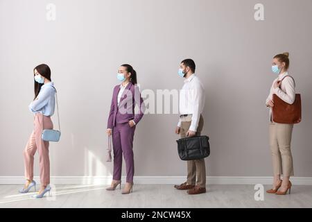 Geschäftsleute mit Schutzmasken warten in der Schlange neben hellen Wänden in Innenräumen. Soziale Distanzierung während der Covid-19-Pandemie Stockfoto