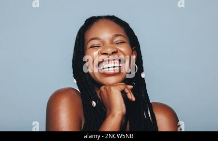 Afrikanische Frau mit Piercings im Gesicht, die fröhlich in einem Studio lächelt. Eine glückliche junge Frau, die Dreadlocks und Piercing-Schmuck gegen eine blaue Hintergrundfarbe trägt Stockfoto