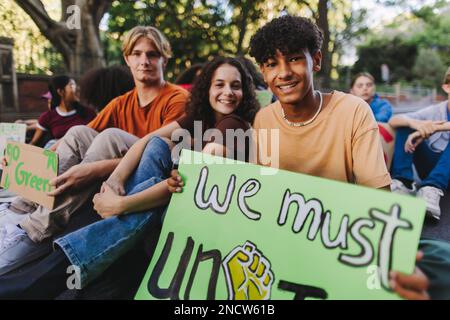 Eine Gruppe multikultureller Aktivisten im Teenageralter, die Poster halten, während sie bei einem Protest gegen den Klimawandel sitzen. Glückliche junge Leute, die während einer Kugel nach Einheit rufen Stockfoto