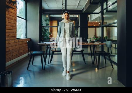 Junge islamische Geschäftsfrau, die in die Kamera schaut, während sie in einem Vorstandssaal steht. Selbstbewusste Unternehmerin trägt einen Hijab in einem modernen Stil Stockfoto