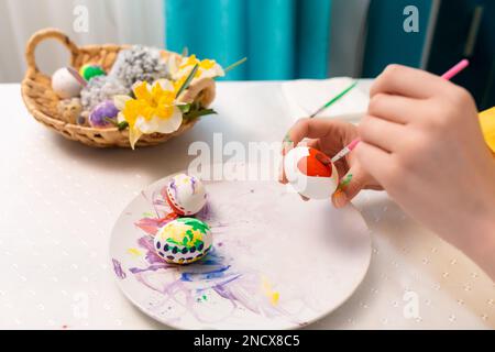 Nahaufnahme von weiblichen Händen, die Ostern Eier dekorieren. Auf dem Tisch steht ein Korb aus Korb mit einem Spielzeughasen und Narzissen. Konzept des traditionellen c Stockfoto