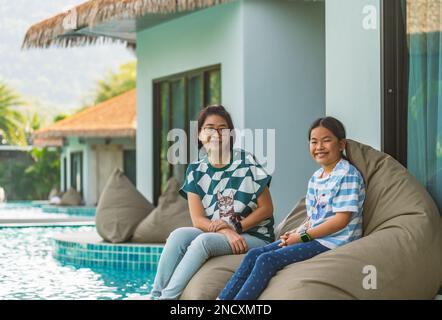 Porträt asiatische Mutter und Tochter sitzen auf einem Sitzsack am Pool eines Resorts, eine Frau mittleren Alters und ein Mädchen mittleren Alters im Alter von 8 oder 9 Jahren. Stockfoto