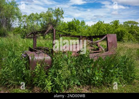 Der rostige Metallrahmen eines antiken Autos befindet sich an einem Sommertag auf einem Feld mit überwuchertem Gras mit Cumuluswolken am Himmel. Stockfoto