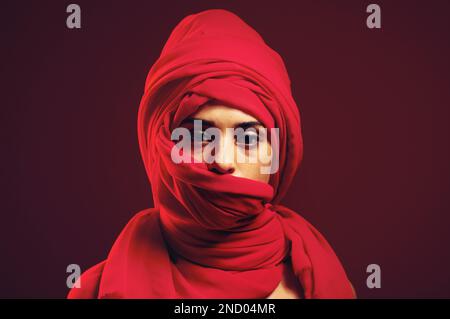 Hidschab, moslem und Porträt einer islamischen Frau im Studio mit dunklem Hintergrund. Ernst, bedeckt und rotes arabisches Kopfkleid mit einer jungen Person Stockfoto