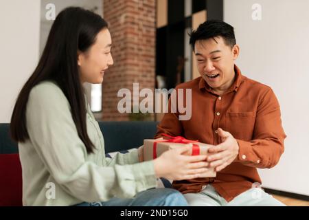 Liebevolle junge asiatische Frau, die ihren Mann mit dem Hochzeitstag begrüßt, ihm eine Geschenkbox schenkt und lächelt Stockfoto