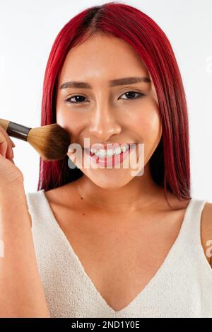 Glückliche junge asiatische Frau mit rotem Haar, die lächelt und in die Kamera schaut, während sie Puder auf das Gesicht mit einem Pinsel auf weißem Hintergrund aufträgt Stockfoto
