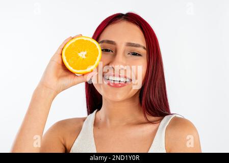 Glückliche junge Frau mit leuchtend rotem Haar, die lächelt, während sie Hälften von Orangen vor weißem Hintergrund hält und in die Kamera schaut Stockfoto
