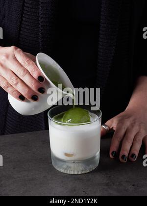 Anonyme Frau gießt aromatisches Matcha-Getränk in einen Glasbecher mit Milch und Eiswürfel während der Zubereitung eines gesunden Getränks Stockfoto