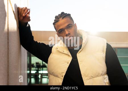 Selbstbewusster junger afroamerikanischer Mann in trendigen Outfits mit Dreadlocks, die auf die Kamera schauen, während sie sich an die Wand des modernen Gebäudes lehnen Stockfoto