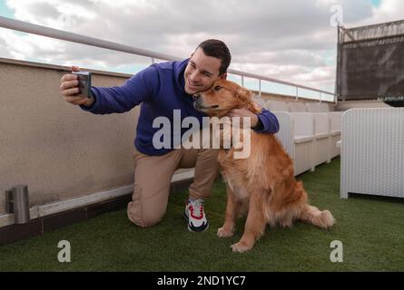 Der ganze Körper eines positiven männlichen Besitzers in legerer Kleidung umarmt den süßen Golden Retriever Hund, während er auf der Terrasse ein Selbstporträt auf einem Smartphone macht Stockfoto