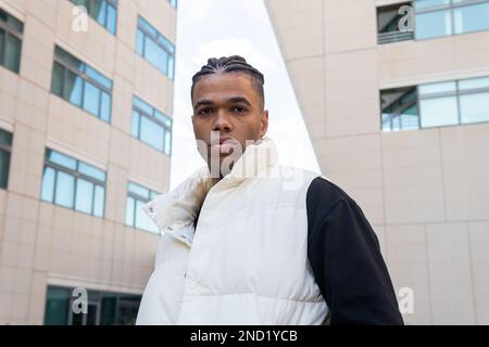 Ein ruhiger junger afroamerikanischer Mann mit geflochtenem Haar in trendigen Outfits, der auf der Straße der Stadt vor der Kamera steht Stockfoto