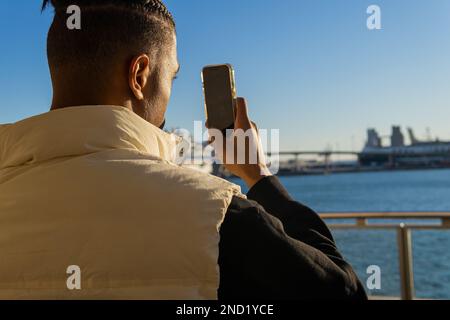 Rückansicht eines konzentrierten afroamerikanischen Mannes mit Dreadlocks, die mit dem Handy Fotos machen, während sie in der Nähe des Ufers stehen Stockfoto