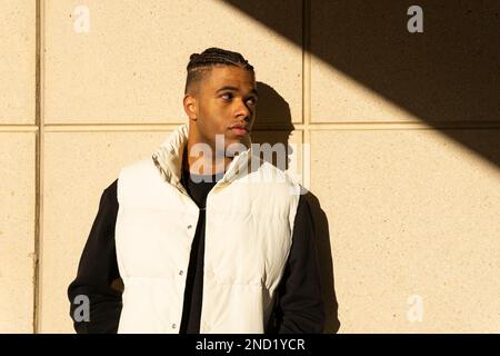 Selbstbewusster junger afroamerikanischer Mann mit geflochtenem Haar in trendigen Outfits, der auf der Straße steht und sich an die Wand lehnt, während er wegschaut Stockfoto