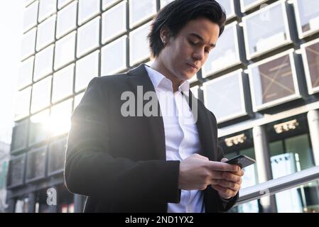 Ein junger Mann in elegantem Outfit, der auf dem Smartphone surft, während er neben einem modernen Gebäude steht und auf den Bildschirm schaut Stockfoto