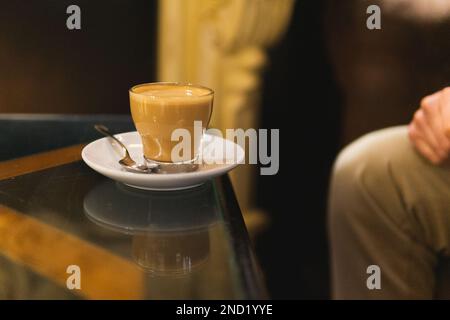 Anonyme Person, die an einem Glastisch sitzt und eine transparente Tasse frischen schwarzen Kaffee mit Sahne im Café serviert Stockfoto