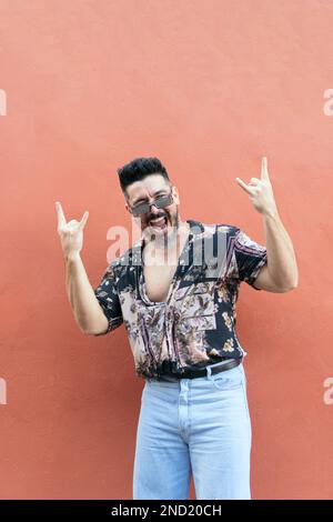 Ein bärtiger männlicher Hipster in einem Sommershirt und eine Jeans mit Sonnenbrille, die mit Felsschildern versehen ist, während er vor orangefarbenem Hintergrund in die Kamera schaut Stockfoto