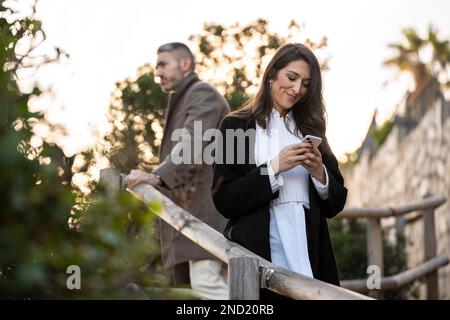 Eine Frau, die auf sein Handy schaut, mit einem wütenden Freund dahinter Stockfoto
