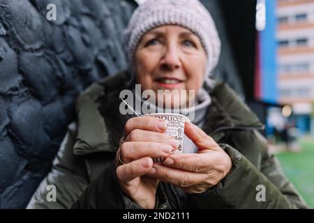 Positive ältere Frau in warmer Oberbekleidung, die eine Tasse heißen Kaffee hält, während sie auf dem Stadionfeld steht Stockfoto