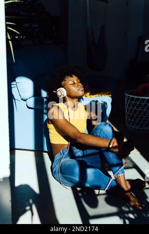 Entspannte junge afroamerikanische Frau in legerer Kleidung, die mit geschlossenen Augen auf dem Boden sitzt und beim Entspannen Musik über Kopfhörer hört Stockfoto