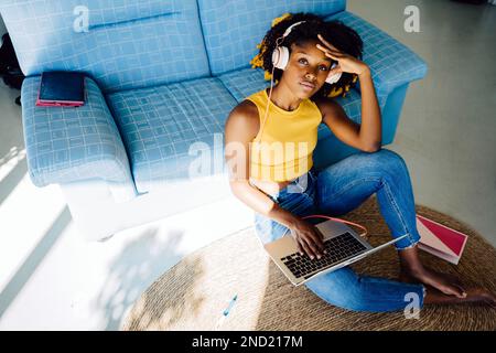 Von oben: Eine nachdenkliche junge afroamerikanische Freiberuflerin in lässigem Outfit, die auf dem Boden sitzt und in die Kamera schaut, während sie in ihrem Notebook surft Stockfoto