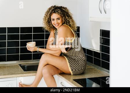 Positive junge, lockige Frau in einem Kleid, die auf der Theke sitzt, während sie zu Hause beim Frühstück Kaffee trinkt Stockfoto